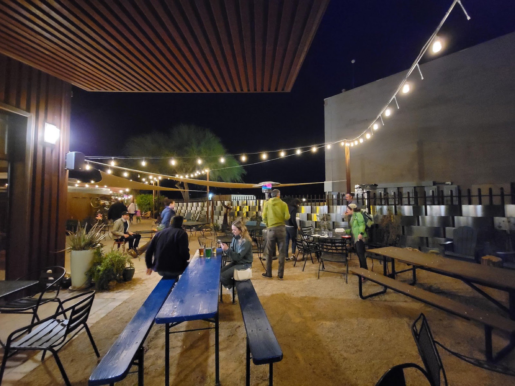 The outdoor beer garden after dark, strung with warm lights over picnic tables and desert plants.