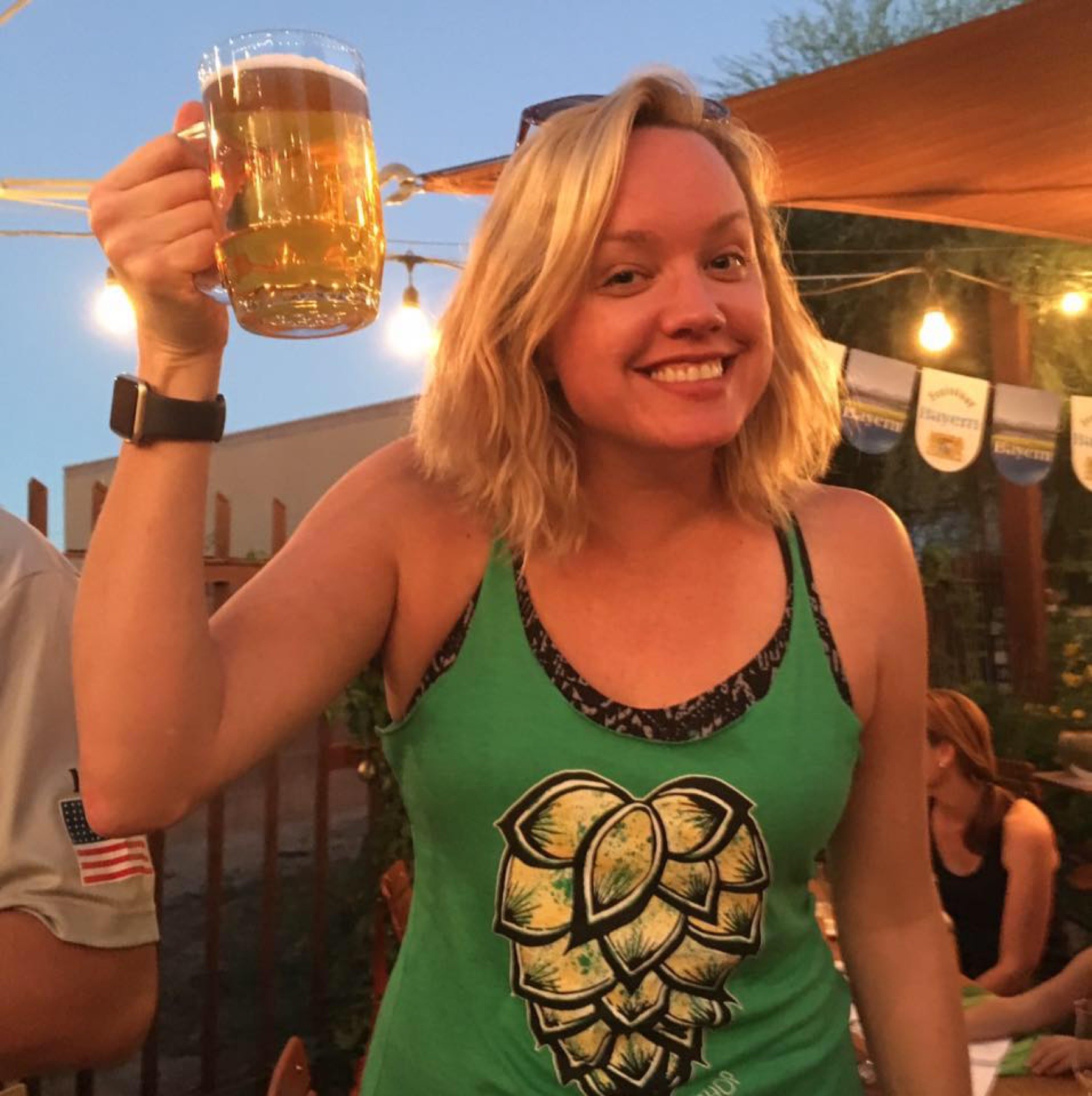 A smiling person raising a frosty mug of beer in the string-lit garden at dusk.